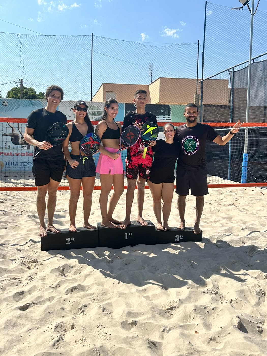 Seis atletas posando no pódio de 1º, 2º e 3º lugares sobre a areia da Ilha Tennis, com raquetes de beach tennis nas mãos