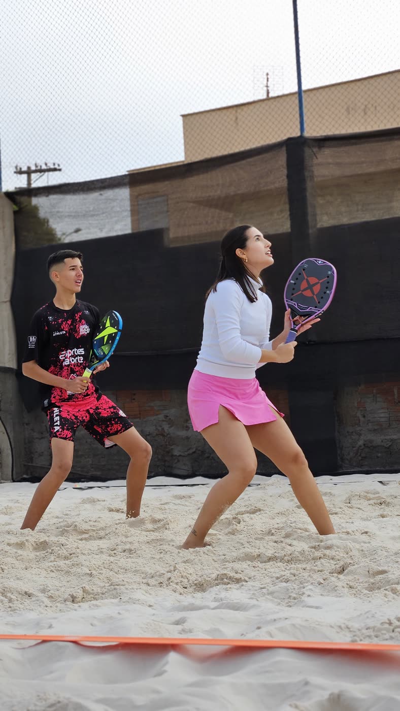 Dupla de beach tennis em quadra no Ilha Tennis, atenta à bola, com raquetes nas mãos e rede de proteção ao fundo